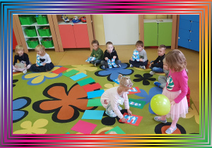 Dzieci siedzą na dywanie. Dziewczynka układa literę na kolorowym kartonie.